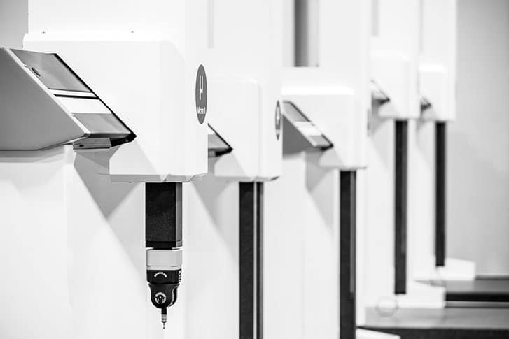 Black-and-white image of multiple coordinate measuring machines (CMMs) in a row, each equipped with a precision probe for detailed measurement, highlighting a clean, industrial environment.